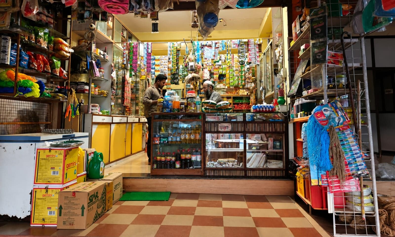 A closer view of the counter inside the shop, with shelves of packaged groceries and two family members attending to it.